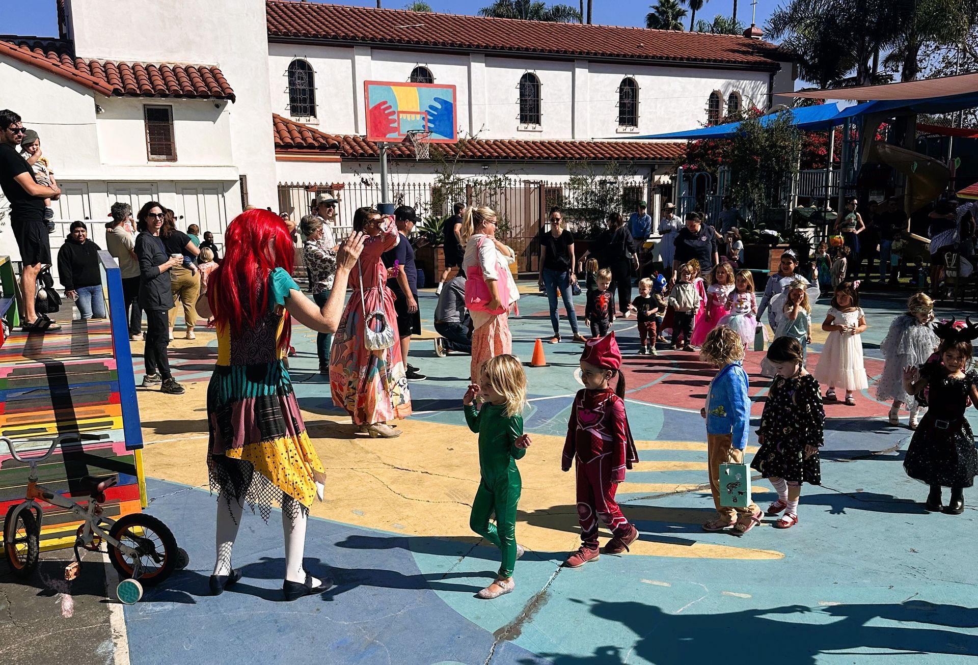 Children in costumes dance on a colorful play area with adults watching. A building is in the background.