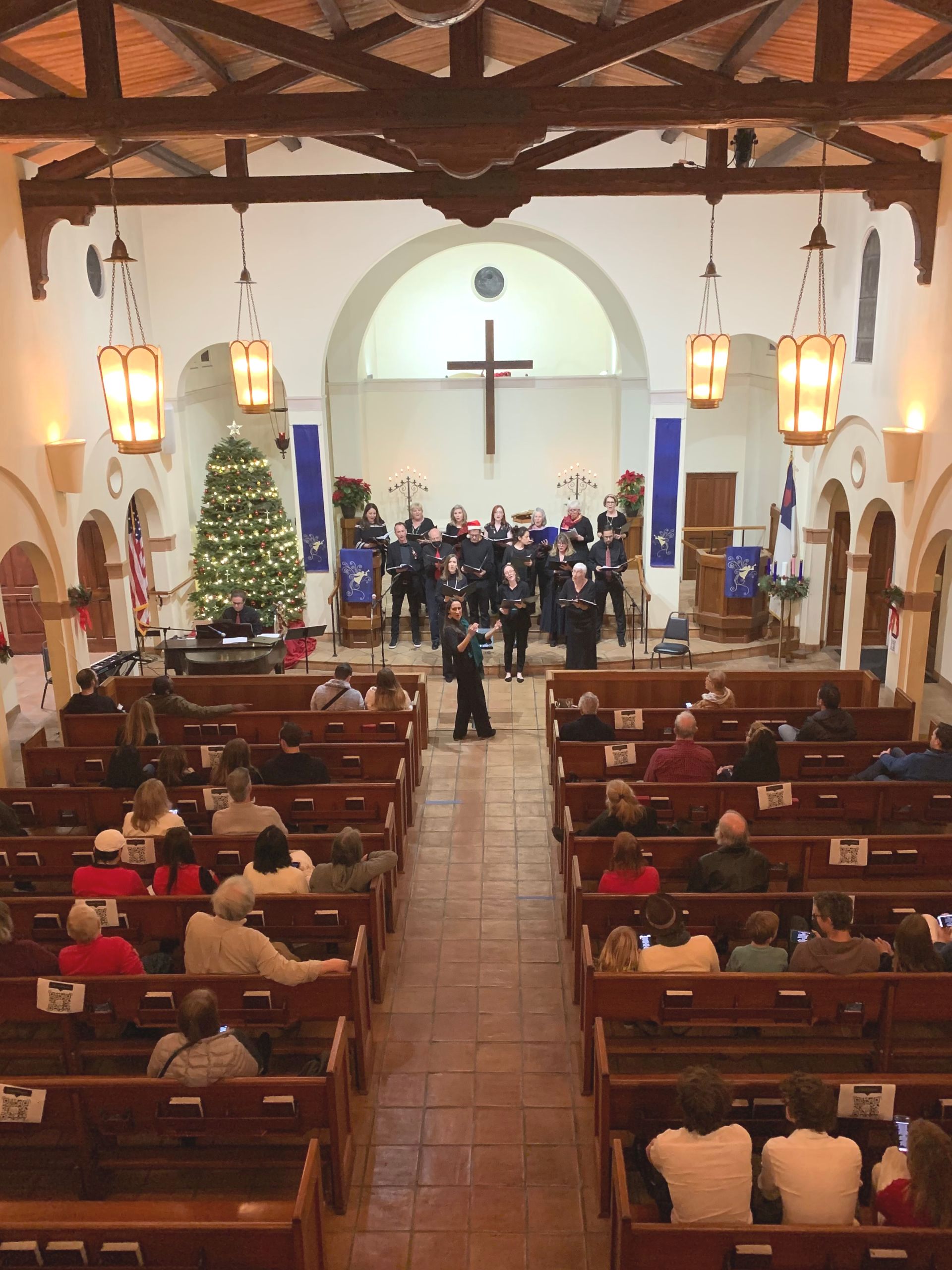 Choir performing in a church with an audience. Christmas tree and cross in the background. Brown pews and walls.