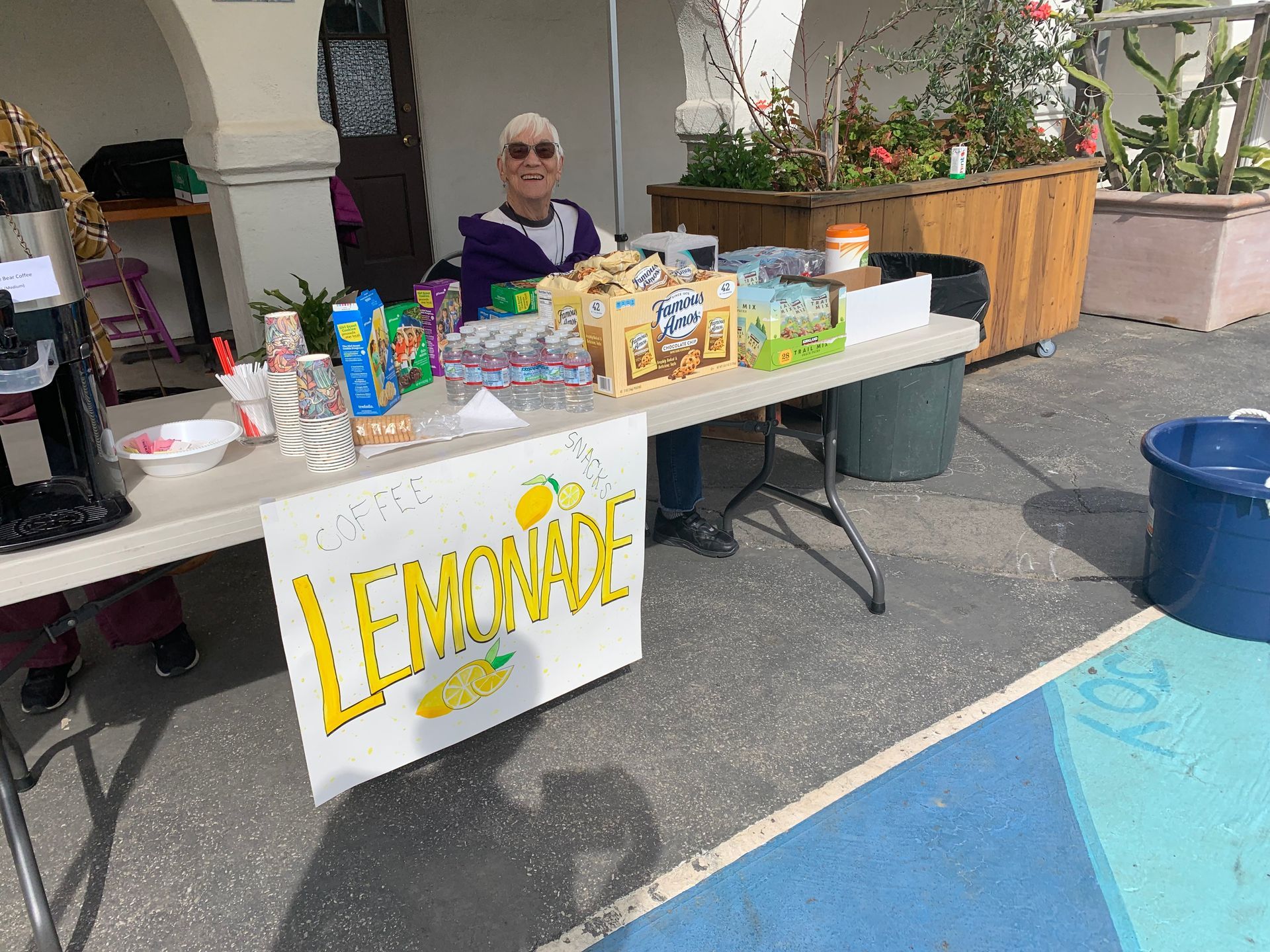 Person at a lemonade stand, selling drinks. The stand has a sign that reads 