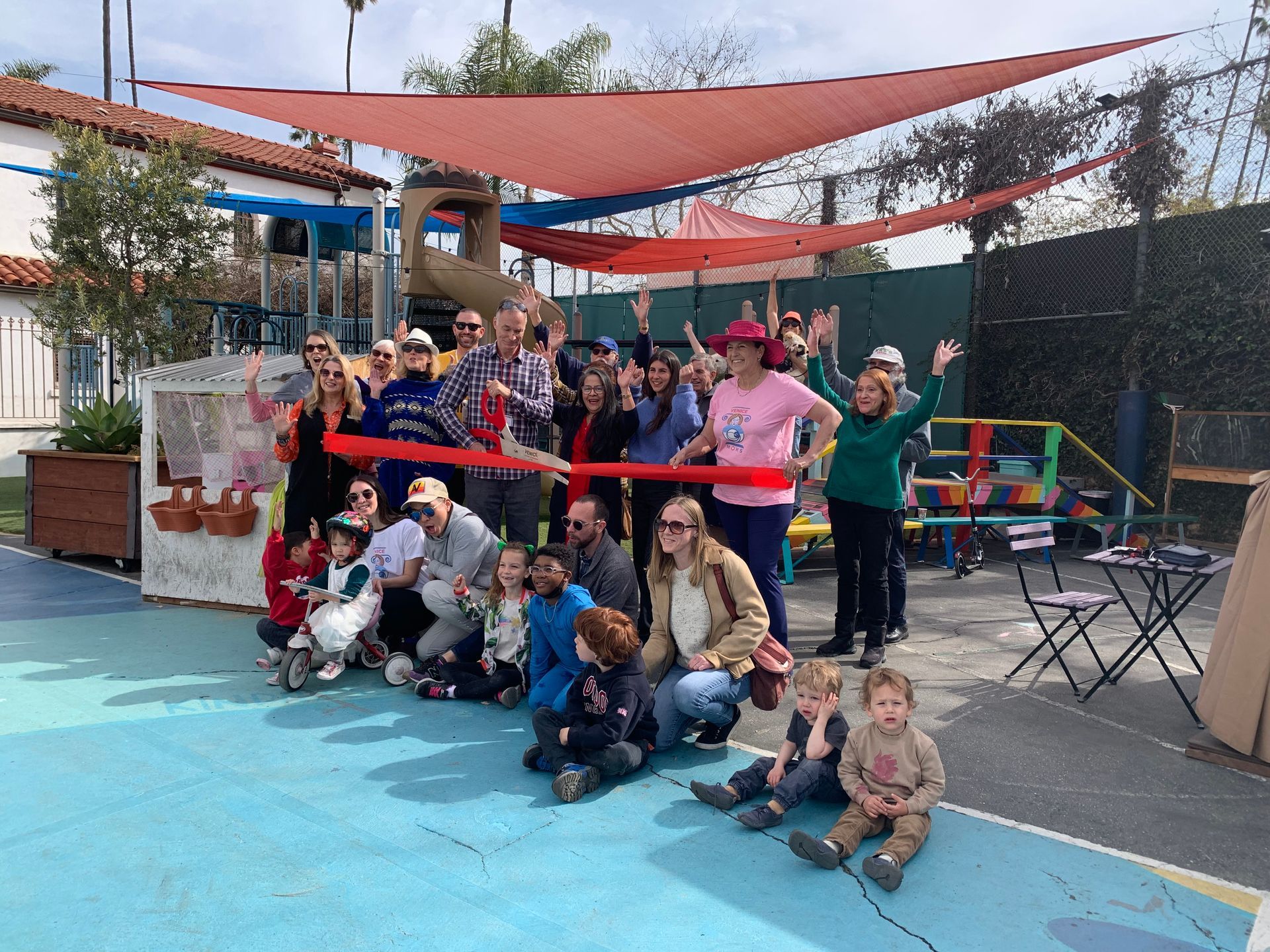 Group of people at a ribbon-cutting ceremony. They are gathered in a playground under a red shade.