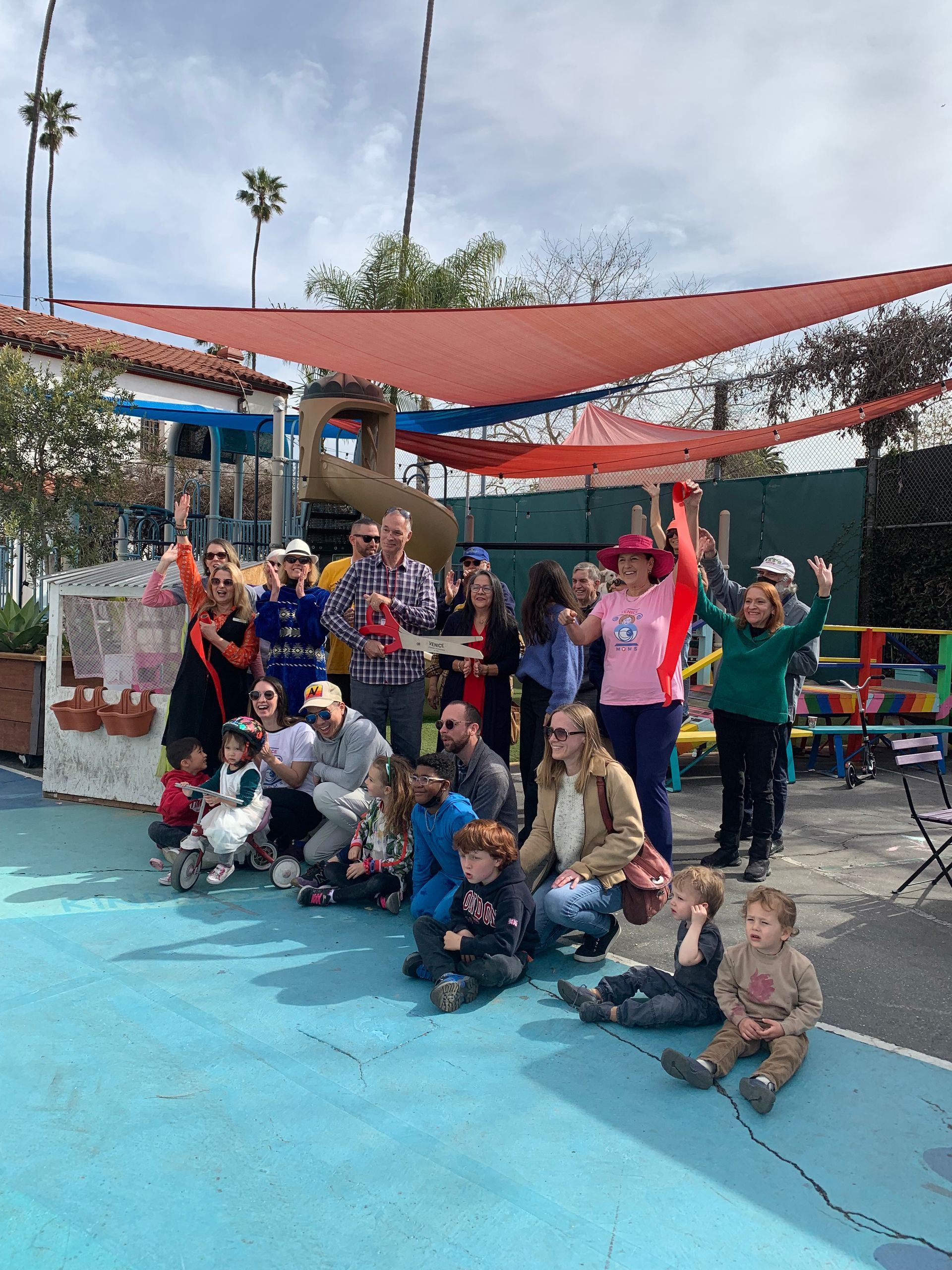 Group of people celebrating outdoors; some holding a red ribbon, others raise hands. Bright blue ground.