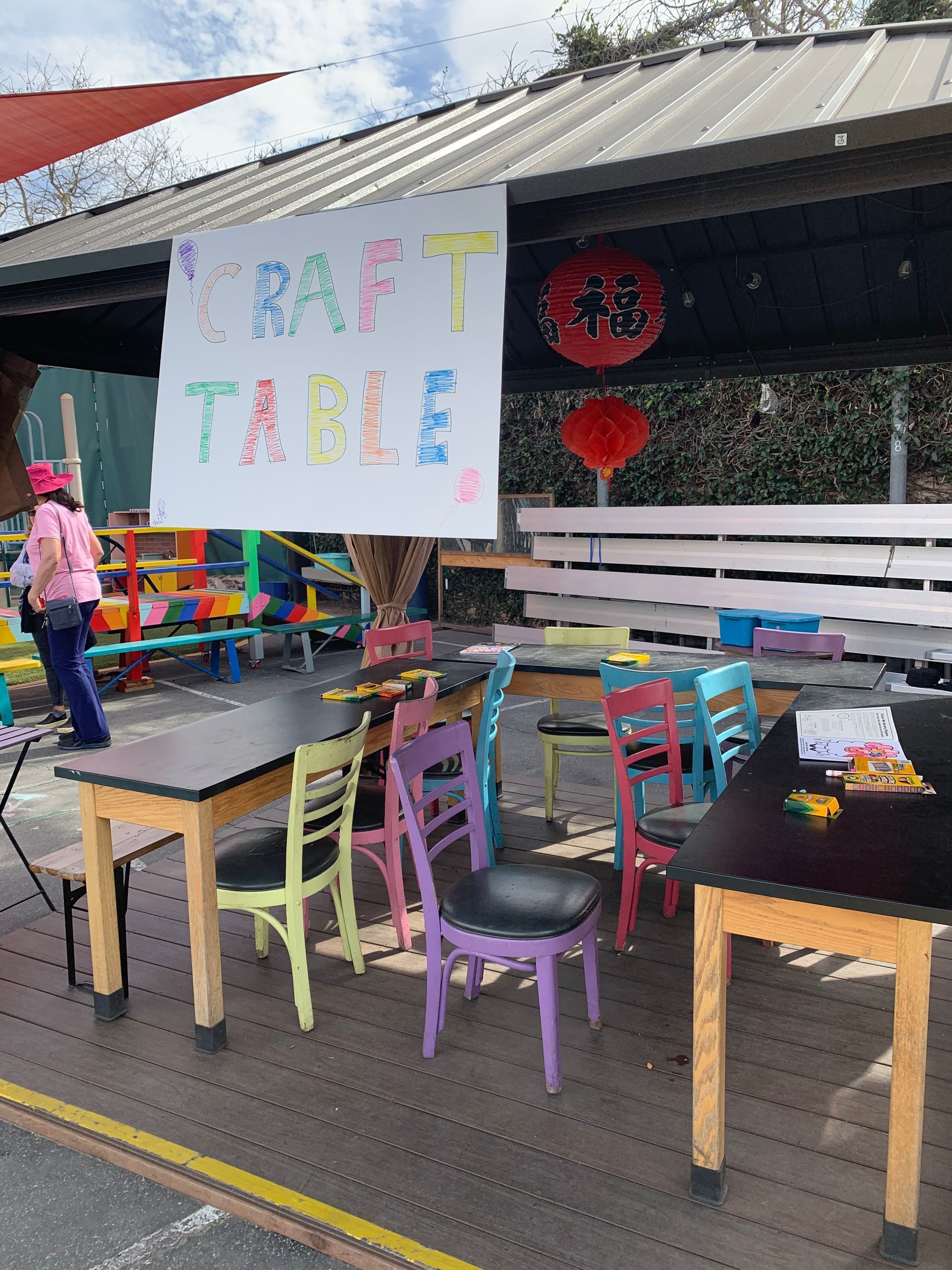 Craft table with colorful chairs under a sign. Someone walks by.