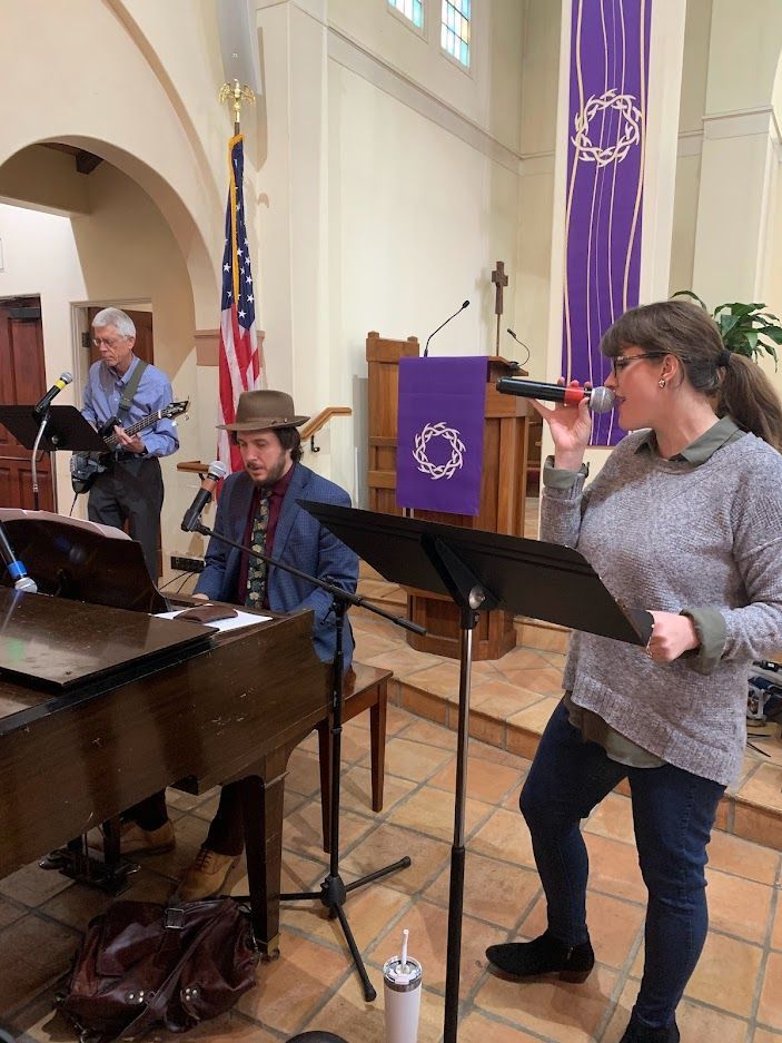 Band performing in church: singer with microphone, pianist, guitarist, and purple banner.