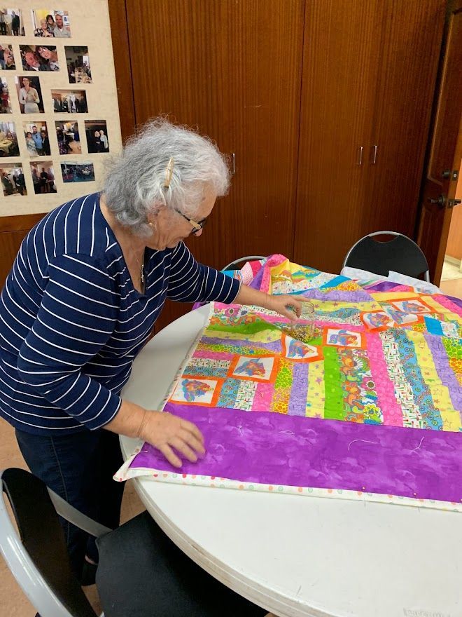 Woman examining a colorful quilt on a round table. Striped shirt, wall of photos, wooden cabinets.