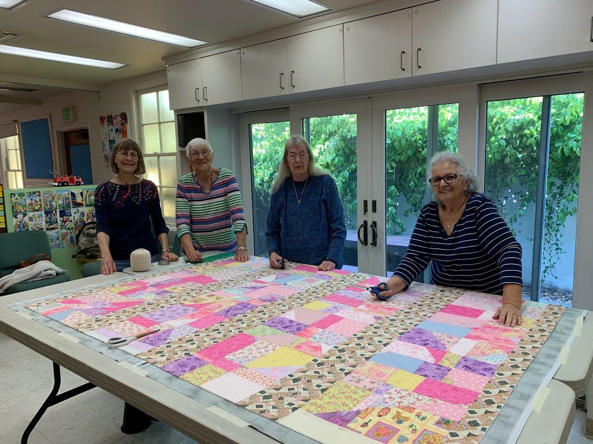 Four women stand around a large, colorful quilt laid on a table in a bright room.