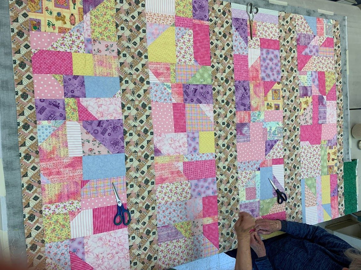 Quilt in progress with pink, purple, and yellow fabrics. Person near scissors and a frame.