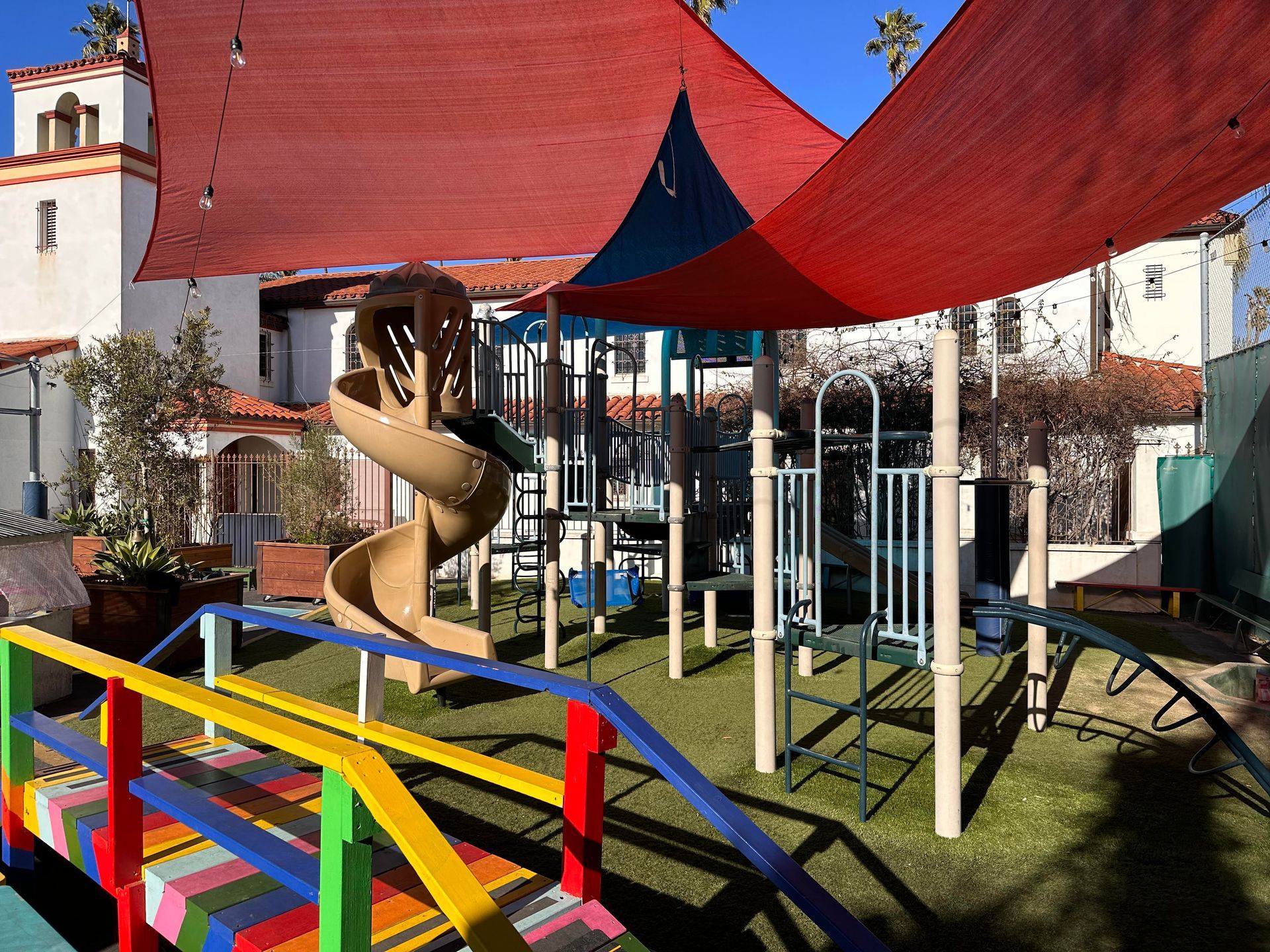 Playground with a slide, bridge, and shade sails, set against a building with a Spanish-style facade.