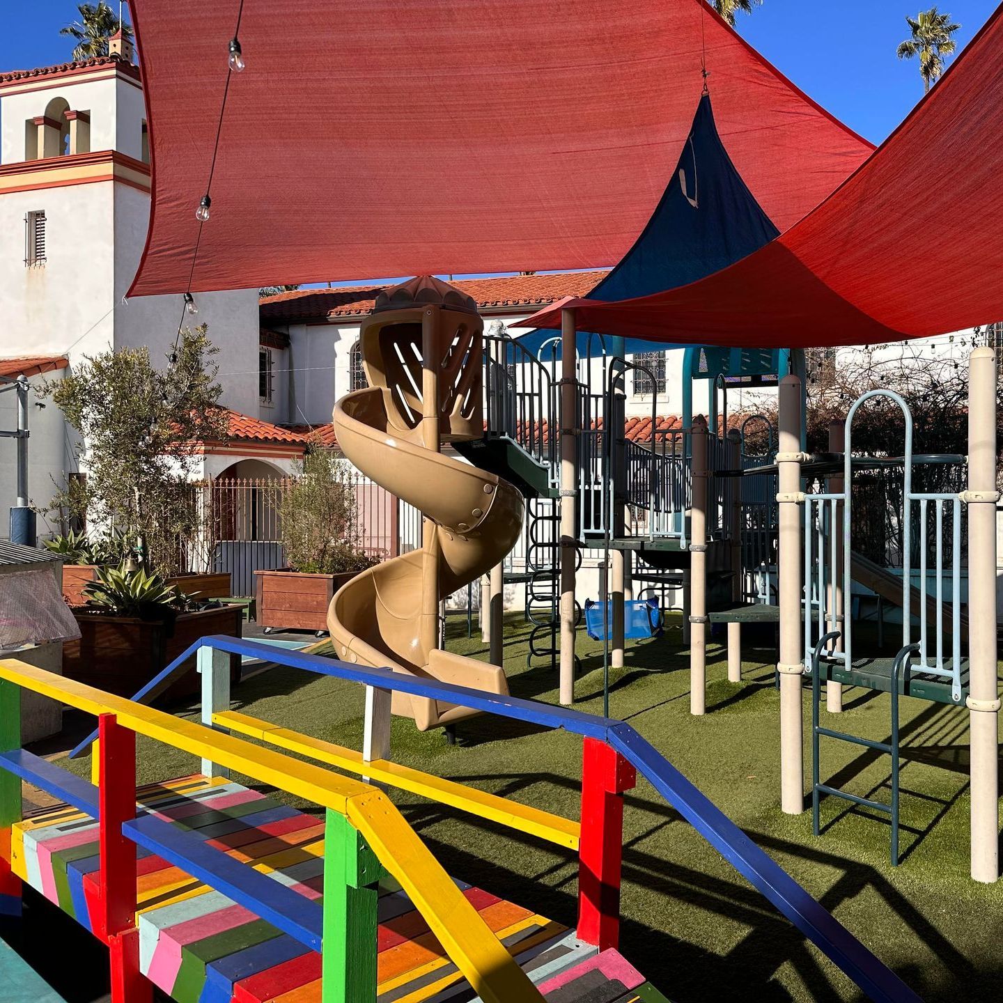Playground with a spiral slide, colorful bridge, and sunshades in front of a white building.