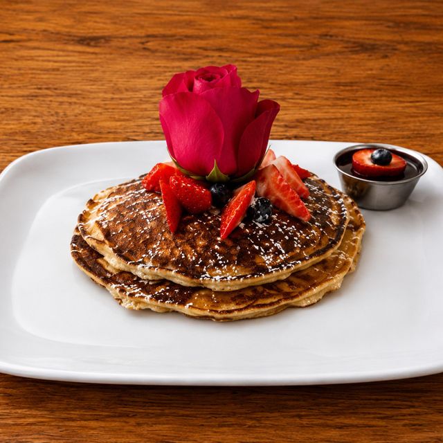 Stack of pancakes topped with strawberries and a red rose on a white plate, with syrup on the side