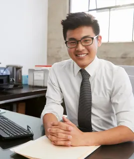 A man in a white shirt and tie is sitting at a desk with his hands folded