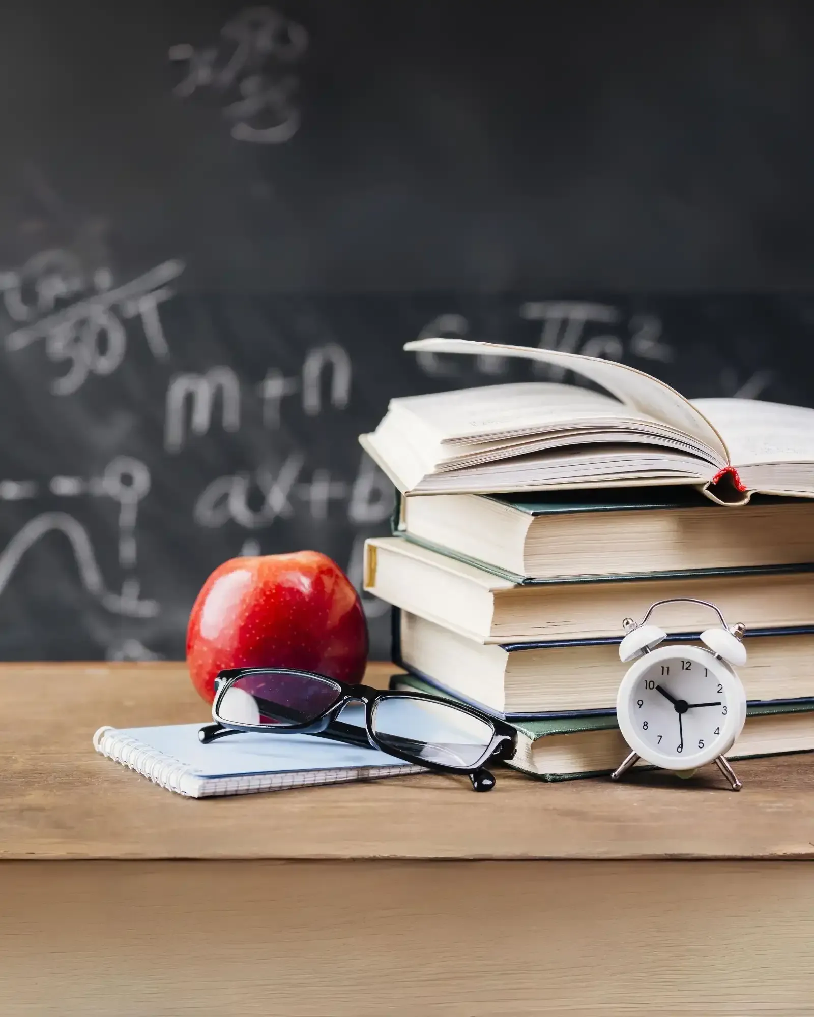 A stack of books , an apple , glasses , and an alarm clock on a desk in front of a blackboard.
