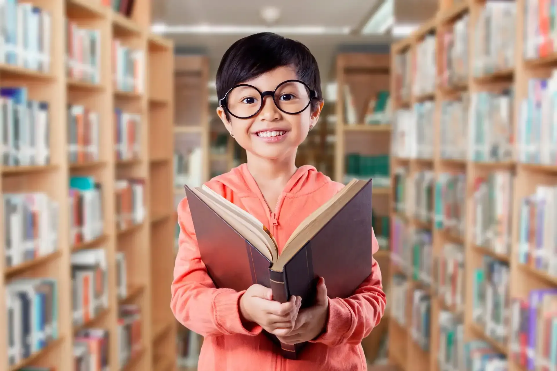 a young girl wearing glasses is holding a book in a library .