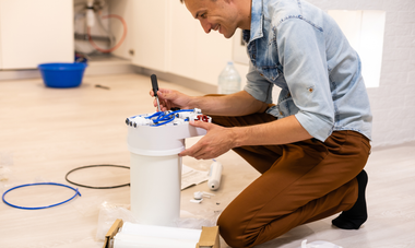 Person kneeling indoors, painting a white bucket with blue paint and a brush on a hardwood floor