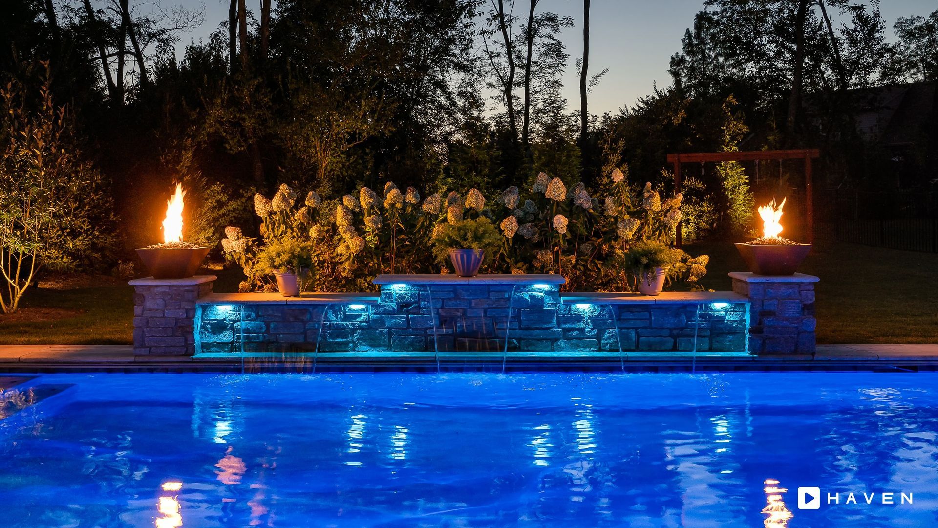 Blue-lit backyard pool with glowing waterfall and torches at dusk