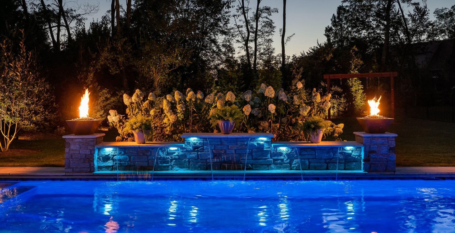 A swimming pool at night with a stone water feature illuminated in blue, flanked by two glowing fire bowls.