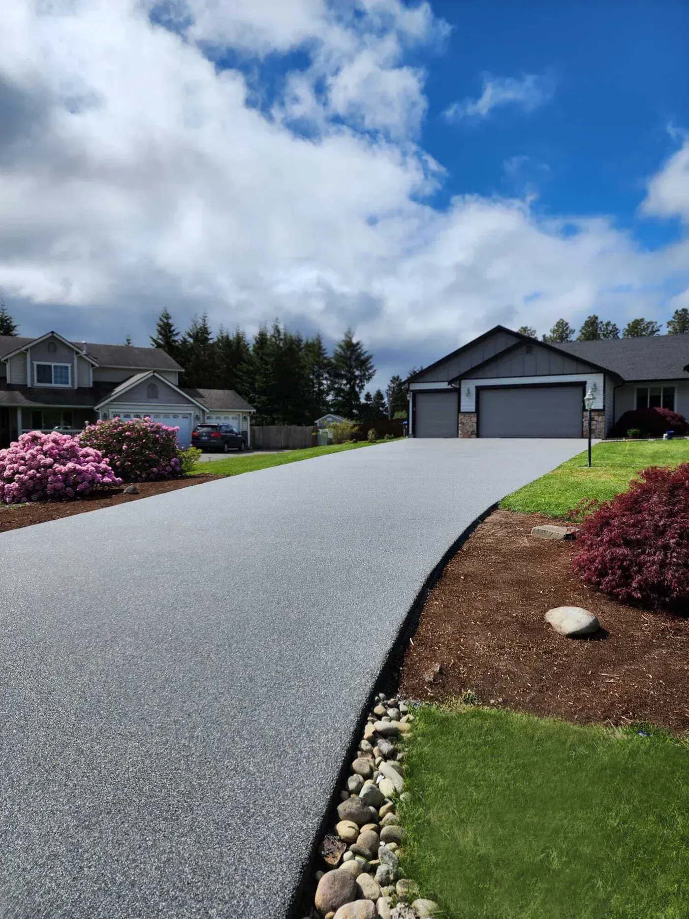 Curved concrete driveway leading to a gray house with garage, landscaped yard, and blue cloudy sky