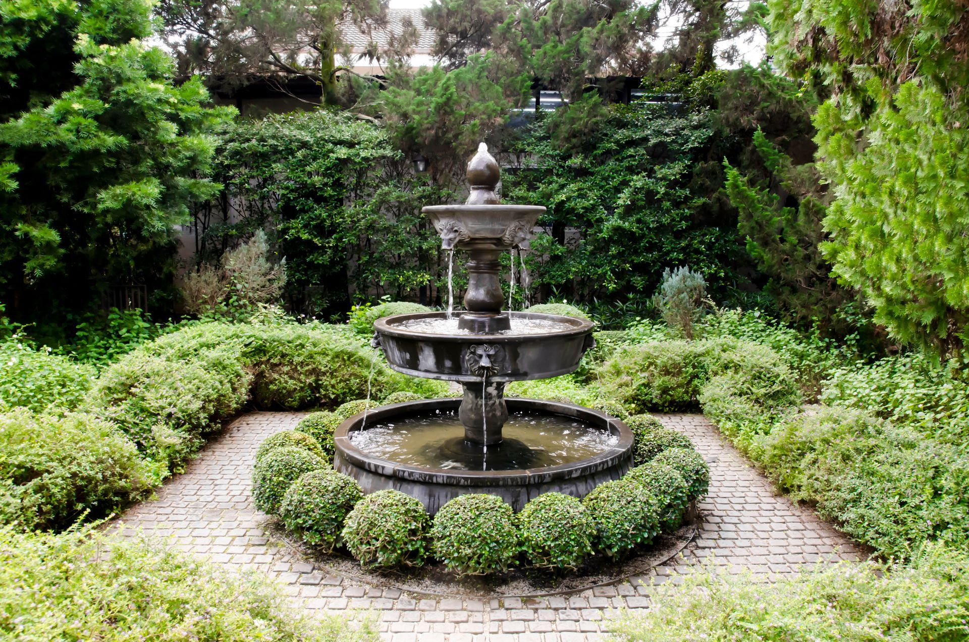Tiered concrete water fountain surrounded by manicured shrubs and brick pavers in a lush garden setting.