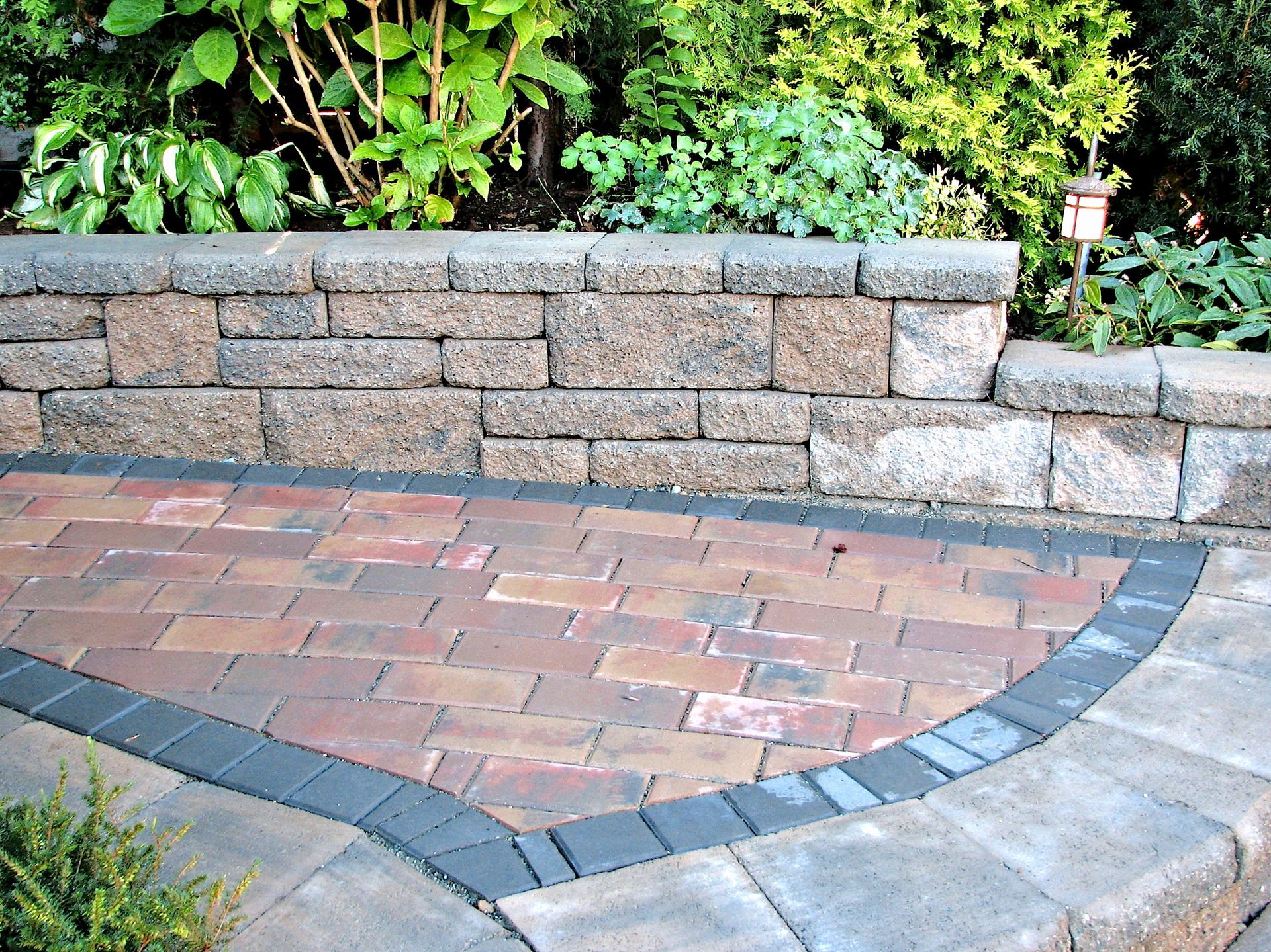 Brick patio with a curved gray paver border beside a stone retaining wall and greenery.