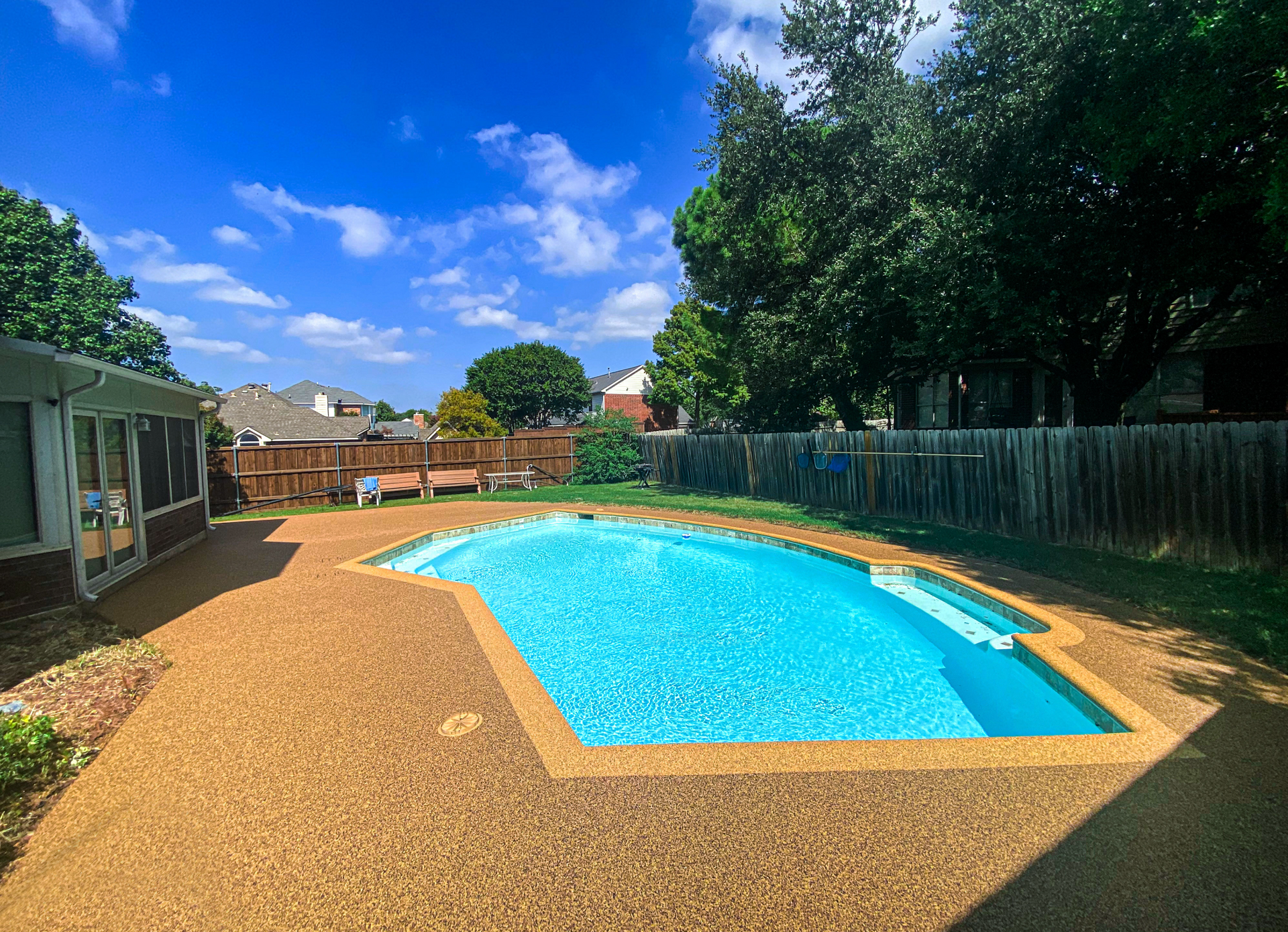 Swimming pool installed by Elite Outdoor Living Solutions in OKC with a blue interior, surrounded by brown concrete and greenery under a blue sky.