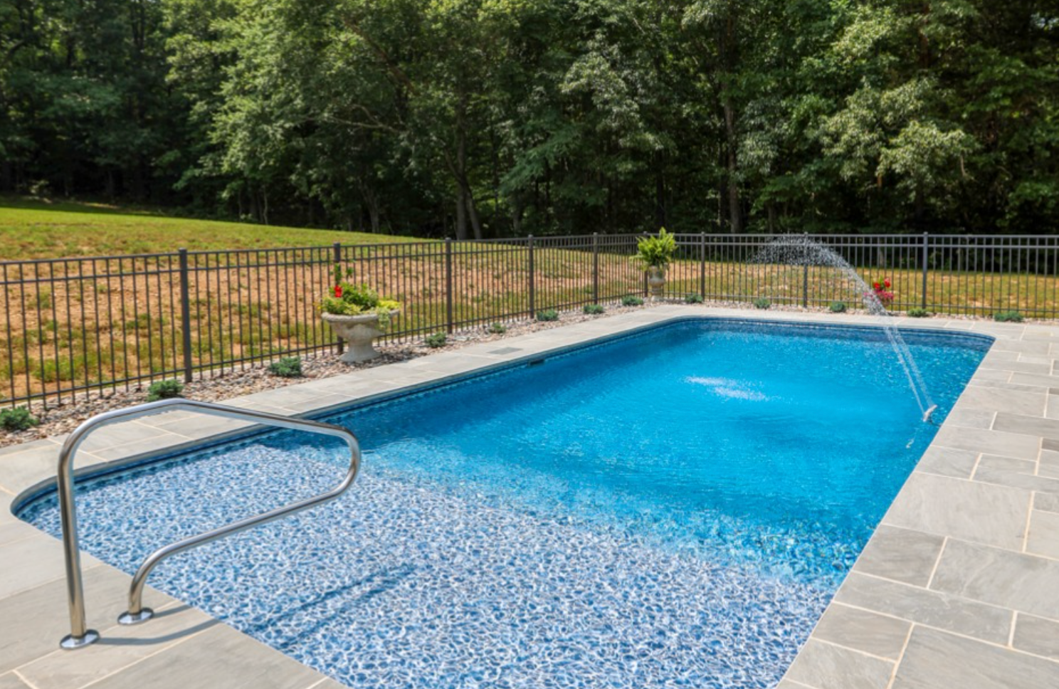 Swimming pool installed by Elite Outdoor Living Solutions in OKC with a blue interior, surrounded by brown concrete and greenery under a blue sky.