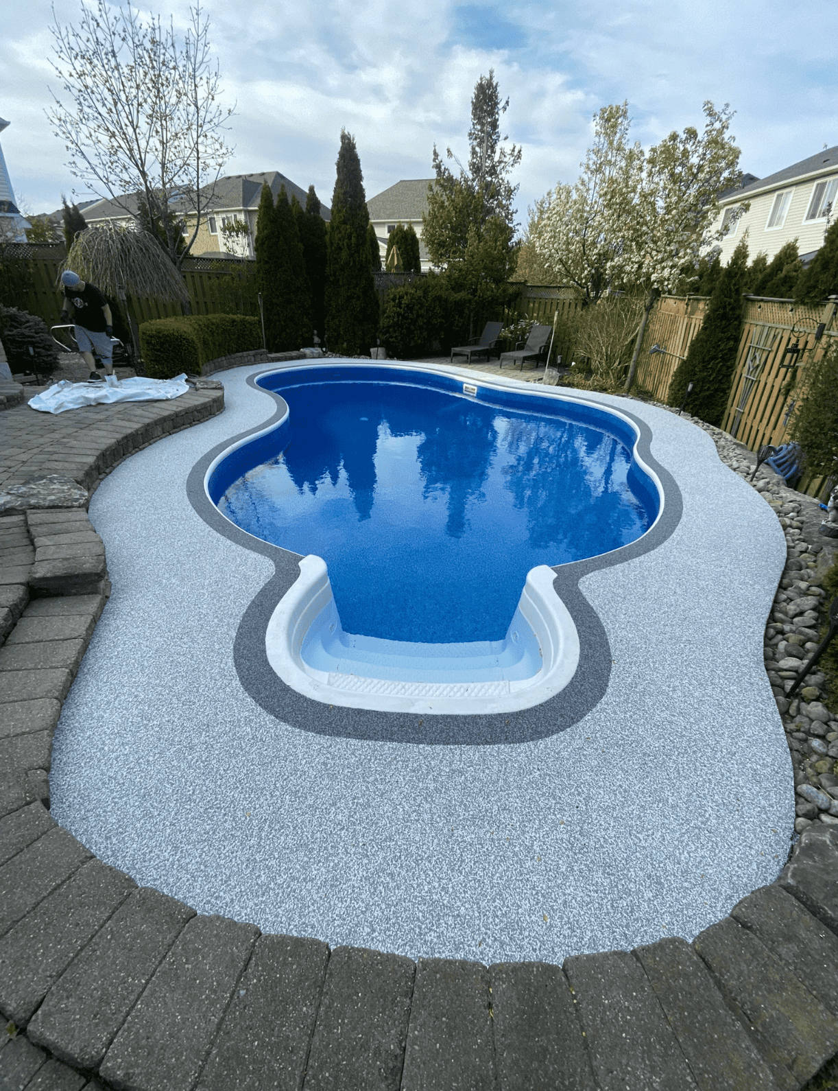 A backyard kidney-shaped swimming pool with blue water and white steps, surrounded by light gray stone aggregate decking.
