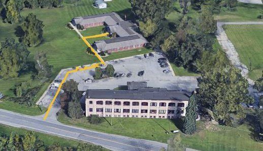 Aerial view of a three-story building, parking lot, and winding driveway beside trees and grass.