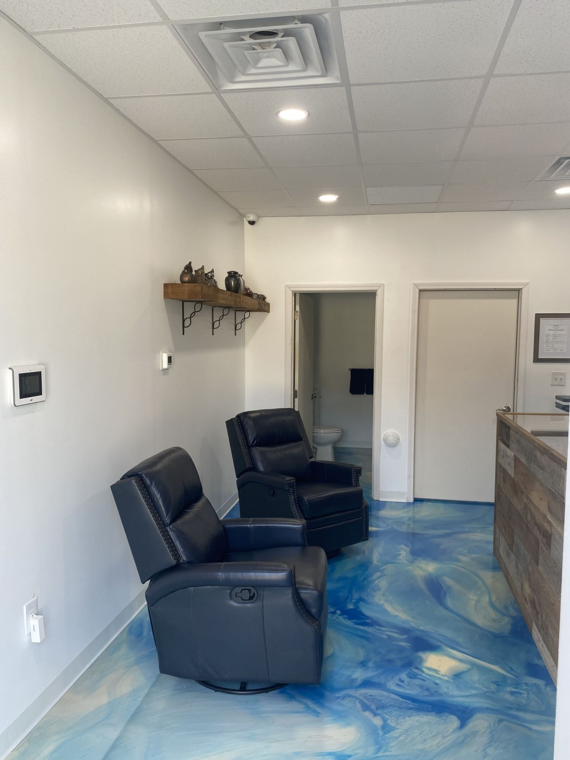 Two black recliners in a waiting area with blue epoxy flooring. White walls, a shelf with objects, and an open doorway.