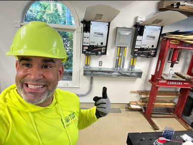 Worker in neon safety gear smiling and giving a thumbs-up in a utility room with electrical panels.