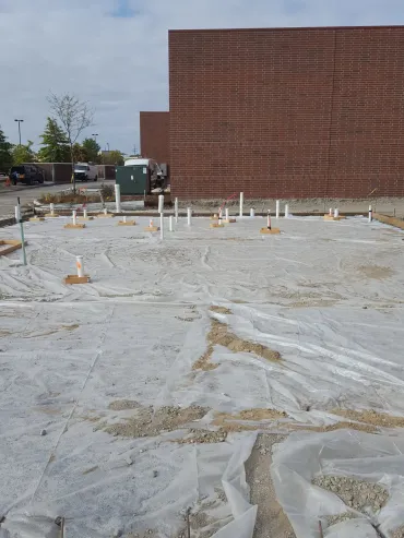 Construction site with concrete foundation, exposed rebar, and a large brick building wall in the background