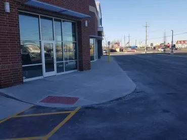 Brick storefront with glass doors, sidewalk ramp, and empty parking lot on a sunny day