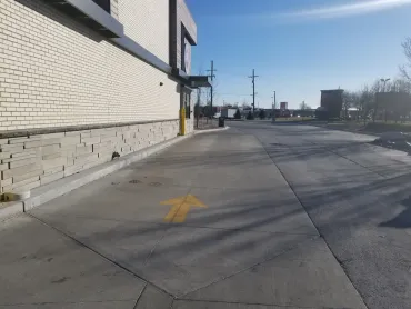 Empty sidewalk beside a white brick building on a sunny day, with a yellow arrow painted on the pavement