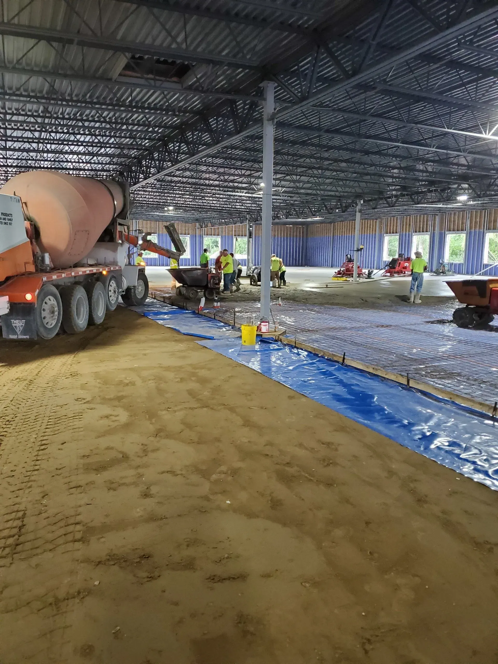 Construction site interior with concrete trucks, steel roof beams, and blue plastic-covered floor under installation