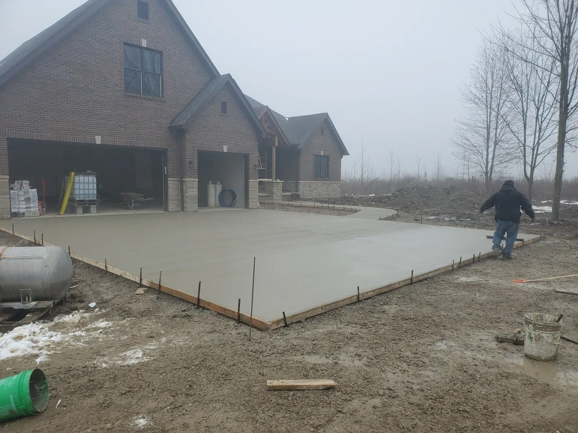 Concrete slab being poured beside a brick house, with a worker smoothing the surface on a foggy day. Concrete slab being poured beside a brick house, with a worker smoothing the surface on a foggy day.