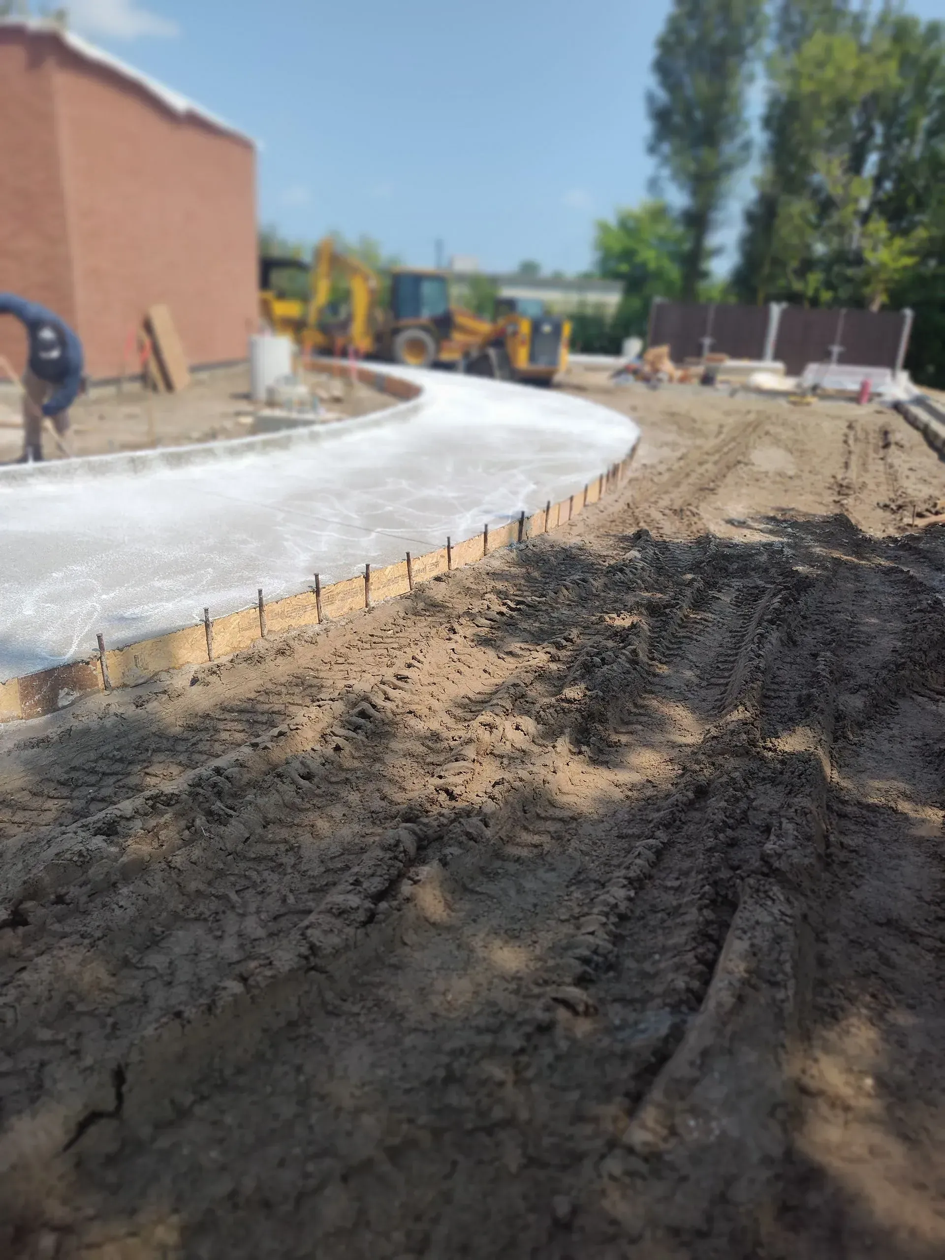Curved concrete path under construction beside a brick building, with dirt, machinery, and workers in the background Curved concrete path under construction beside a brick building, with dirt, machinery, and workers in the background