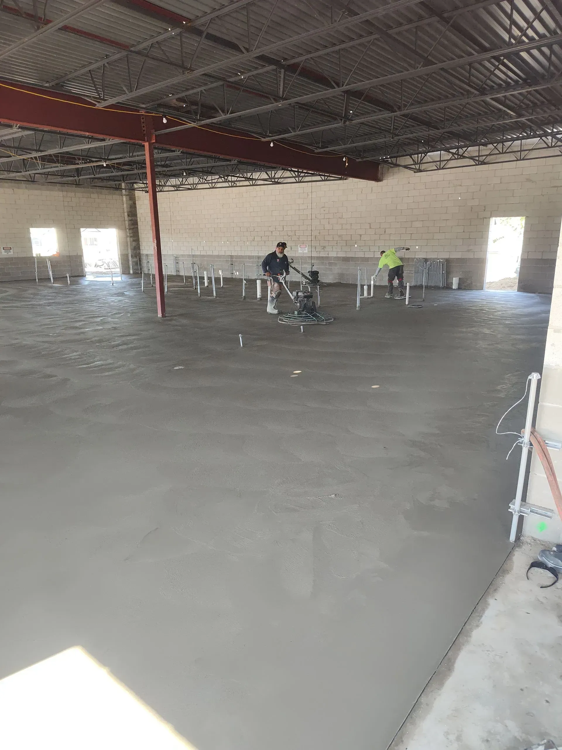 Large unfinished indoor hall with concrete floor, exposed ceiling, white walls, and a few workers in the distance. Large unfinished indoor hall with concrete floor, exposed ceiling, white walls, and a few workers in the distance.