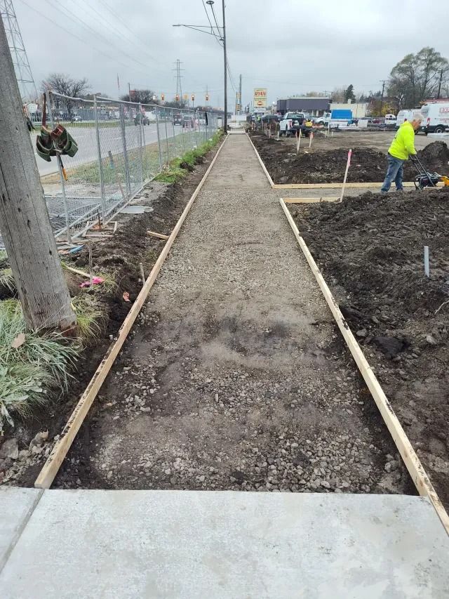 Narrow gravel path beside a fenced roadside garden, with a worker in a yellow jacket on the right. Narrow gravel path beside a fenced roadside garden, with a worker in a yellow jacket on the right.