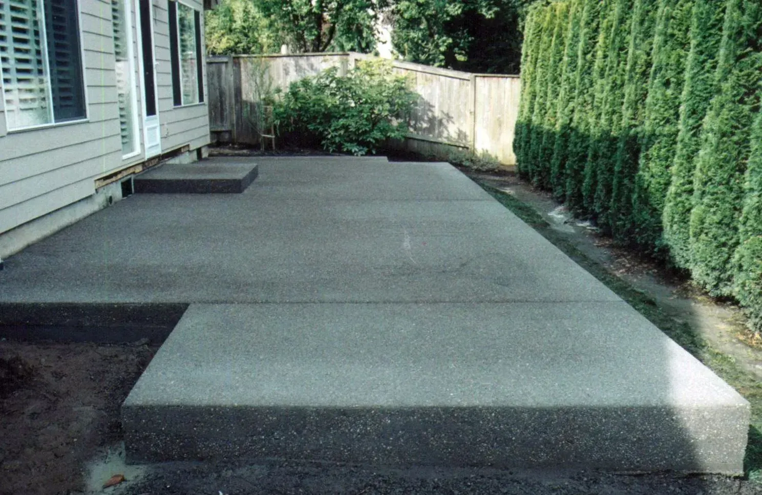 Concrete patio beside a house with raised steps and a tall hedge along the right side. Concrete patio beside a house with raised steps and a tall hedge along the right side.