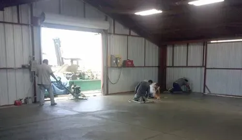 Inside a large metal workshop, two people work near the open doorway with a ladder and tools. Inside a large metal workshop, two people work near the open doorway with a ladder and tools.