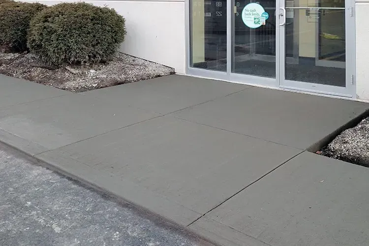 Concrete sidewalk leading to a glass storefront entrance with shrubs on the left and right Concrete sidewalk leading to a glass storefront entrance with shrubs on the left and right