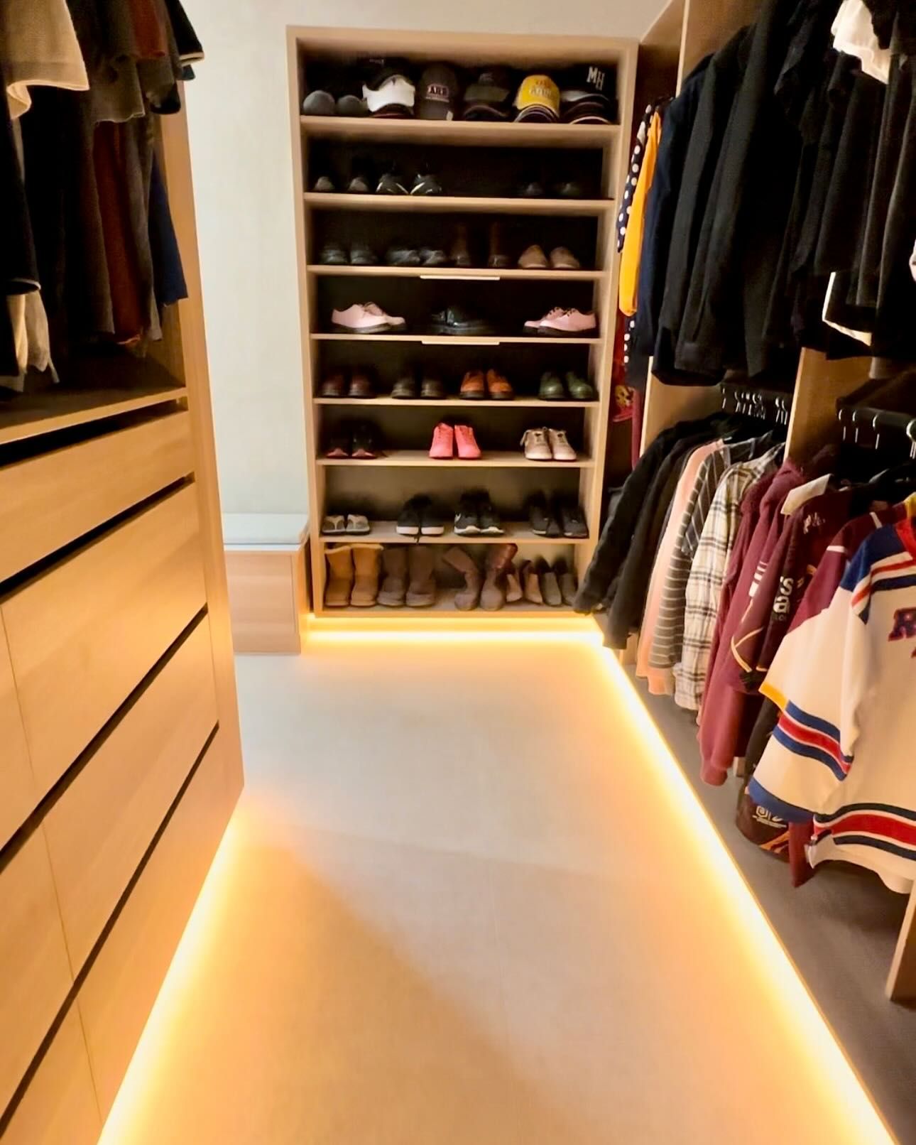 Walk-in closet with lit floor, shoes on shelves, and hanging clothes. Wooden cabinets and neutral carpet.