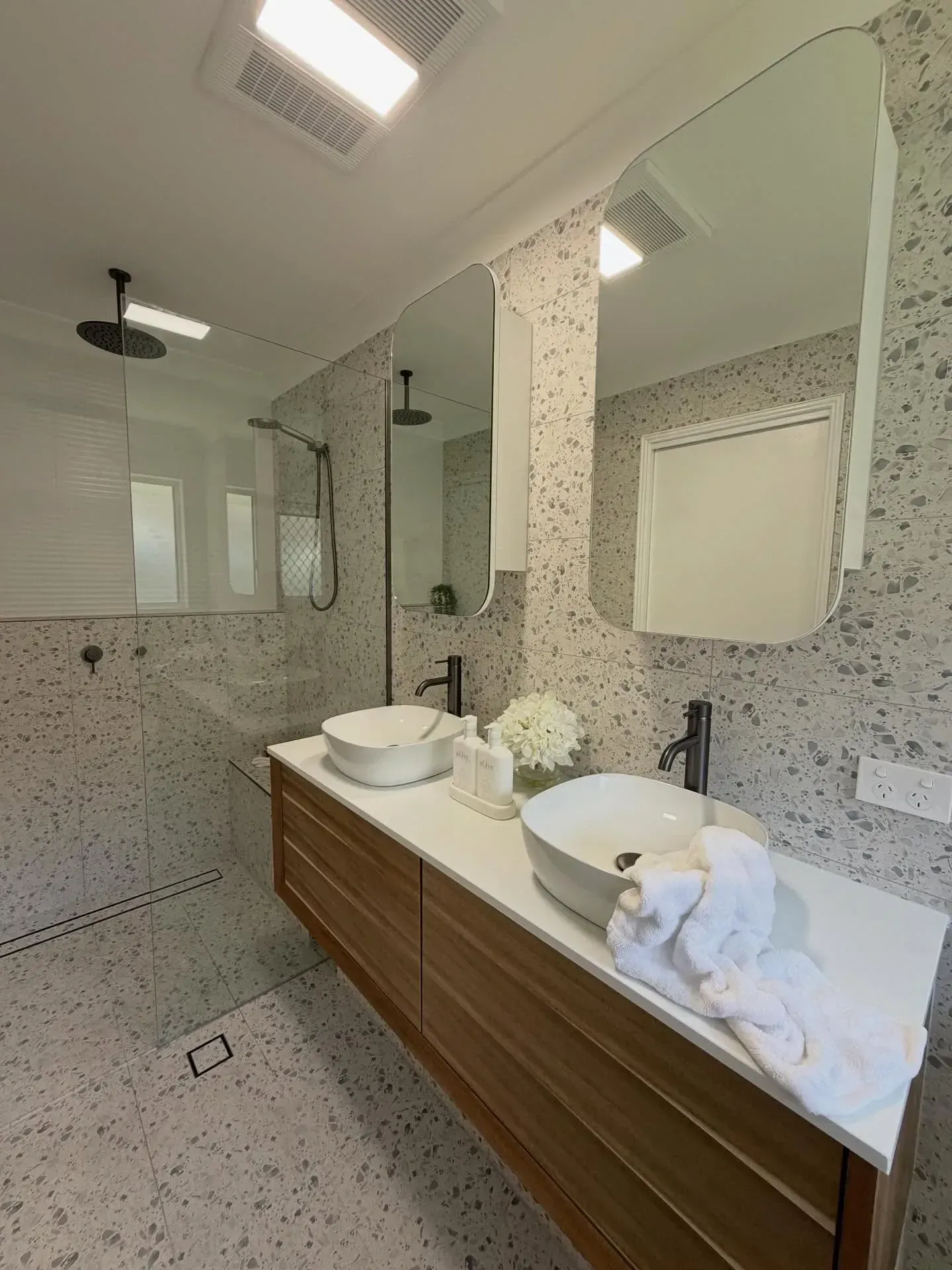 Modern bathroom with double sink, white countertop, speckled wall, and wood cabinet.