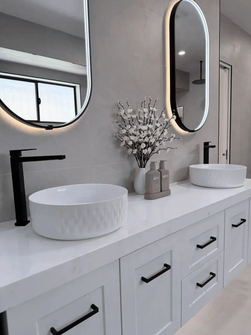 Modern bathroom with white sinks, countertops, and cabinets. Black faucets, oval mirrors, and flower arrangement.
