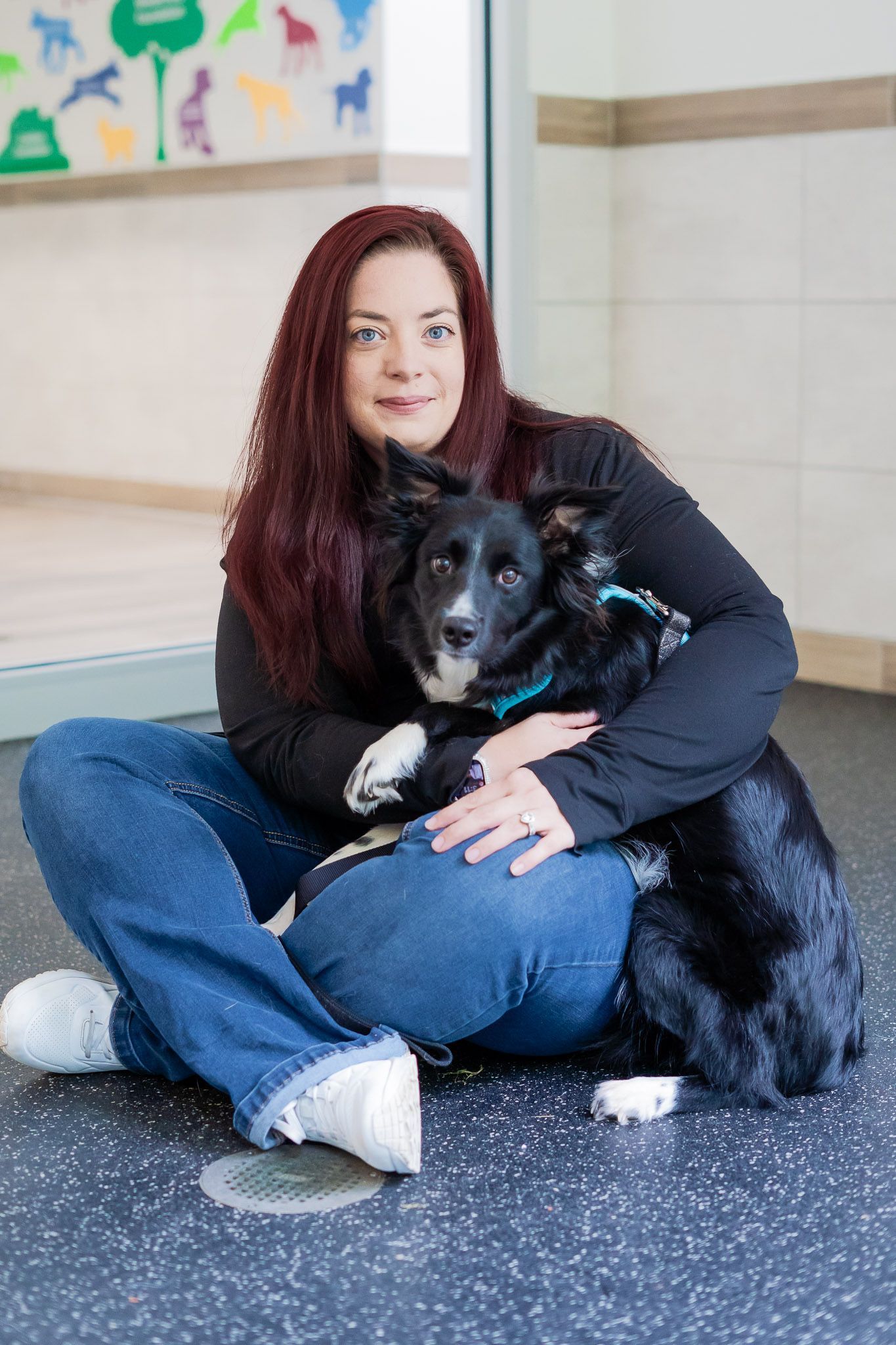 A woman is sitting on the floor hugging a black dog.