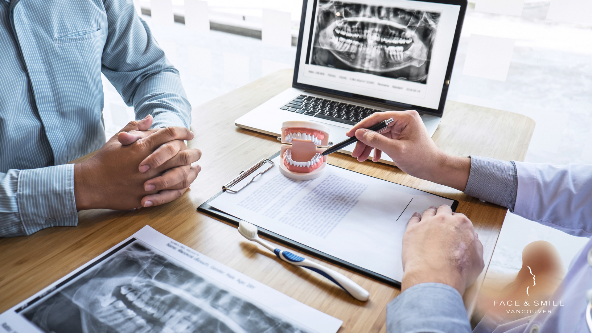 A dentist is talking to a patient at a table while looking at an x-ray on a laptop.
