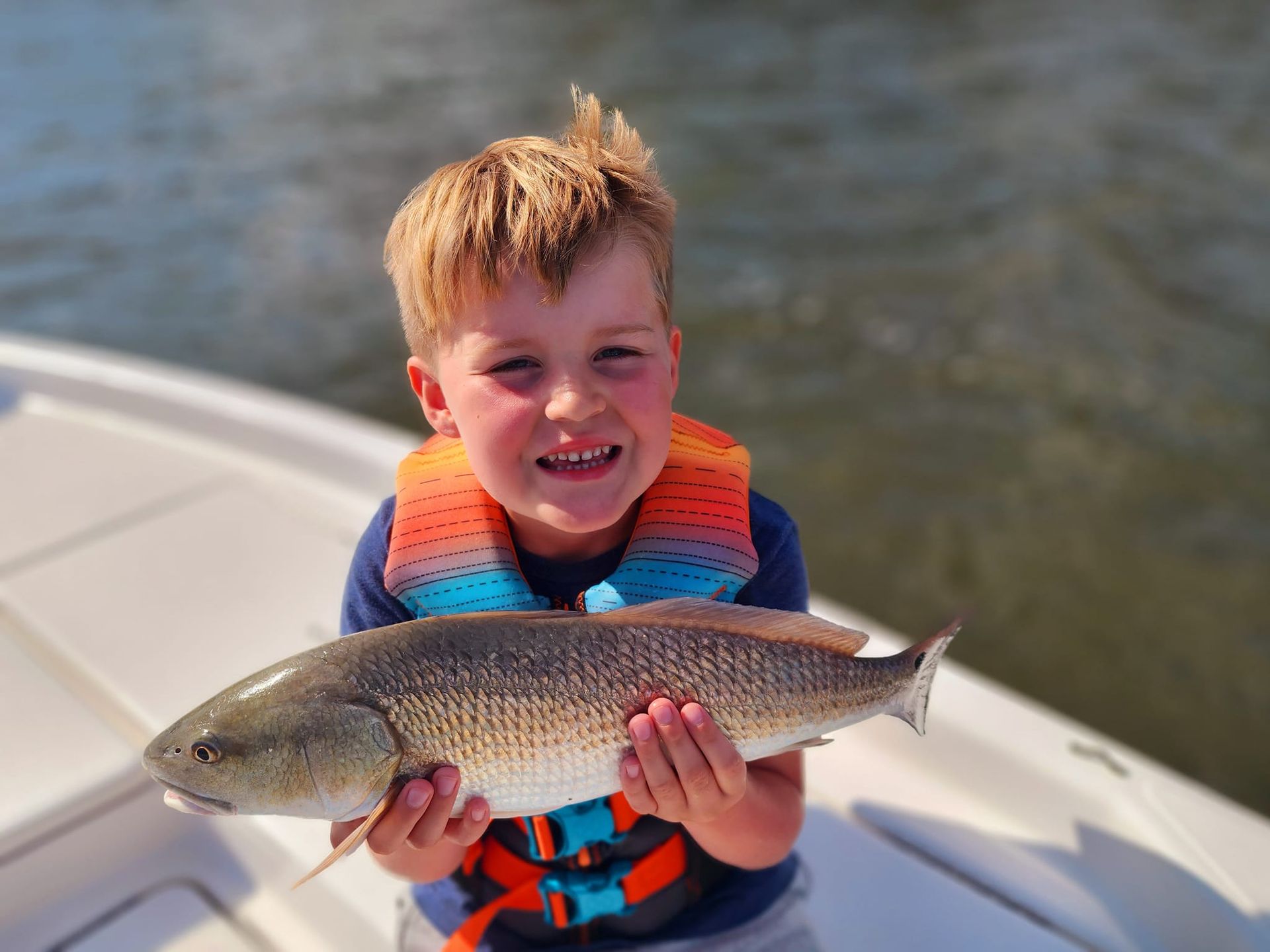 Young child on a boat smiles while holding a reddish-brown fish. He wears a life jacket.