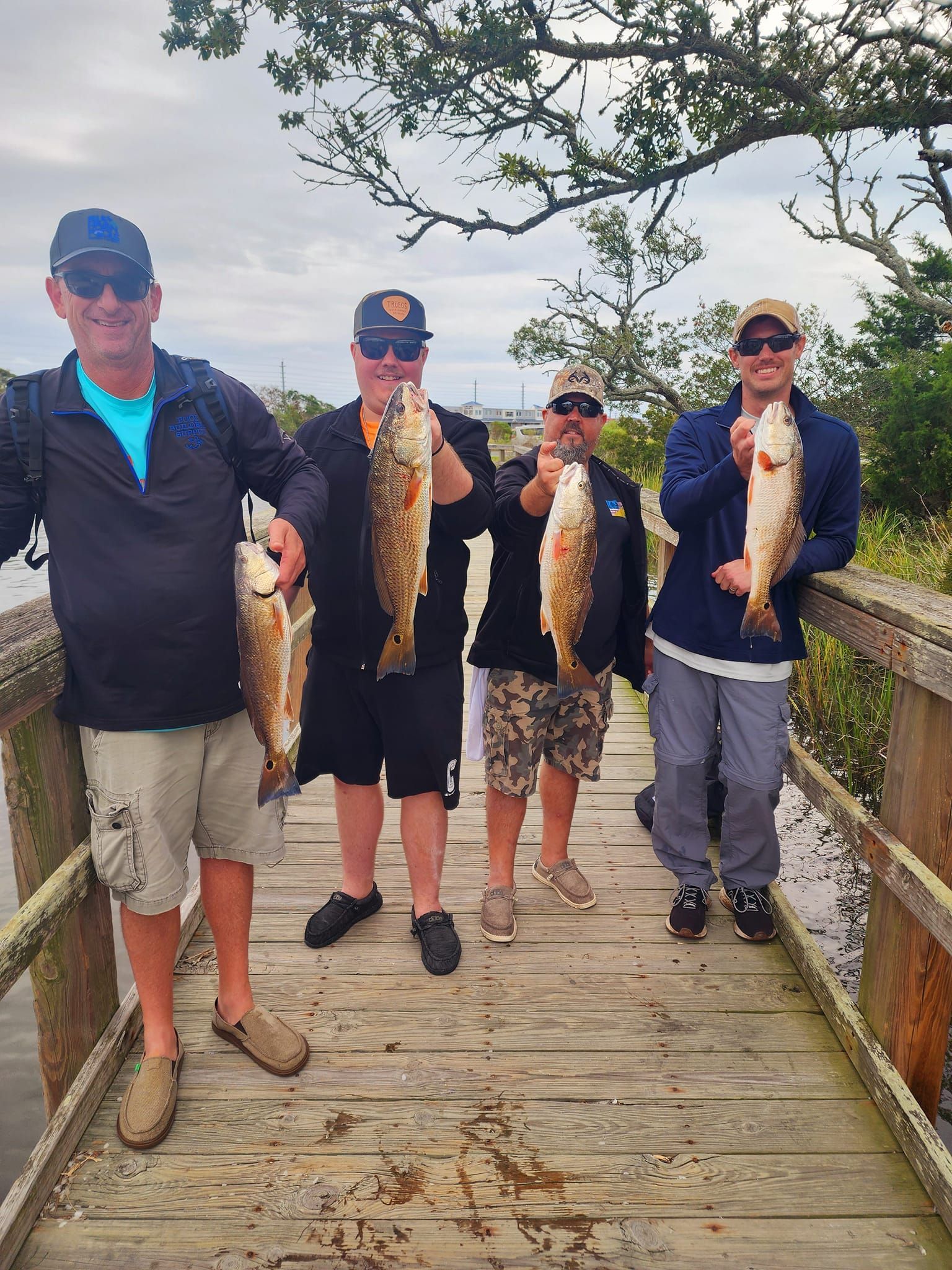 Four people on a wooden dock holding up caught fish. Cloudy sky, trees in the background.