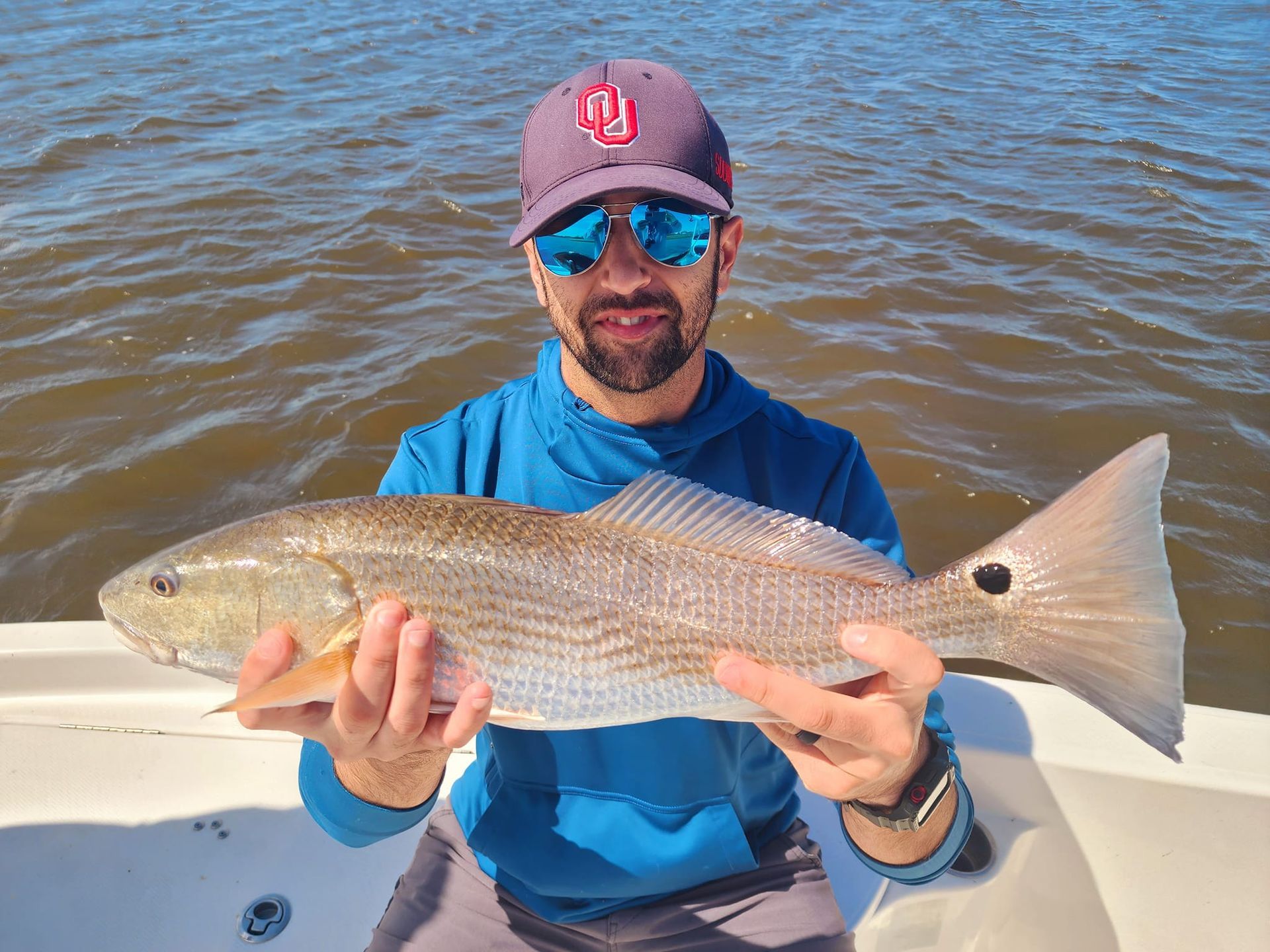 Man holding a large redfish on a boat in sunny water. He's wearing a blue hoodie, sunglasses, and a baseball cap.