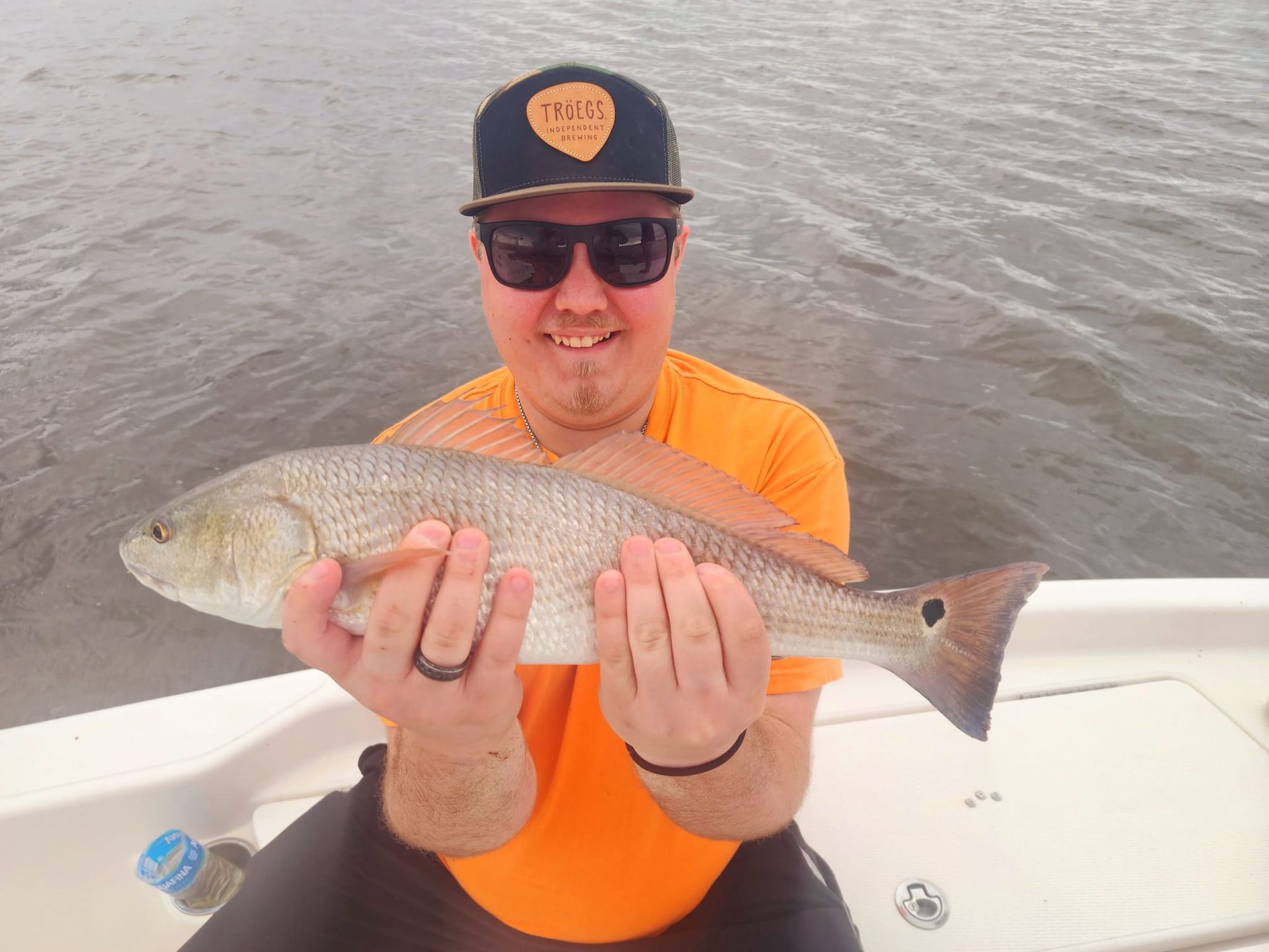 Man in orange shirt and sunglasses holding a redfish on a boat, smiling.