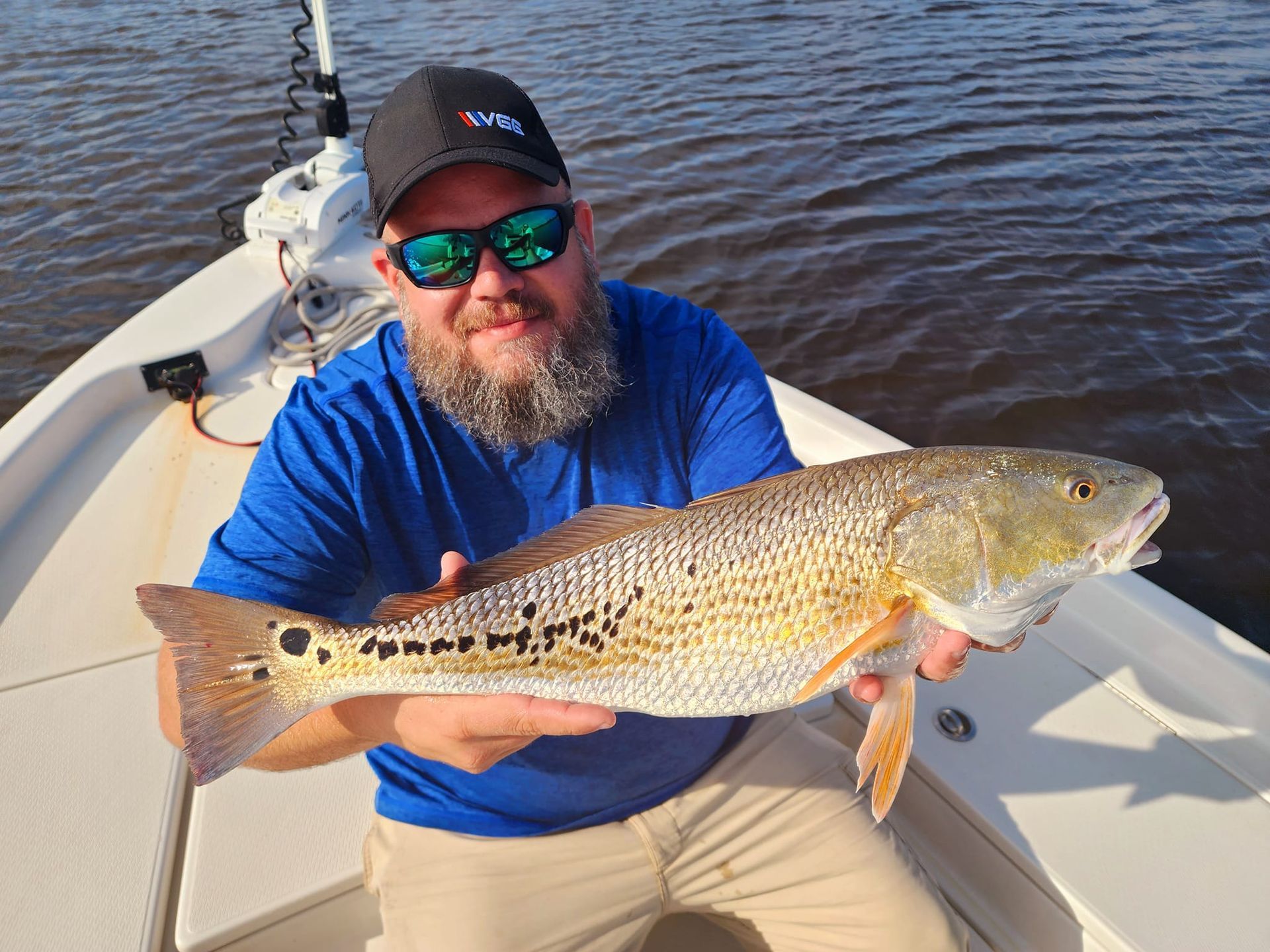 Man in sunglasses and hat on a boat holding a large redfish.