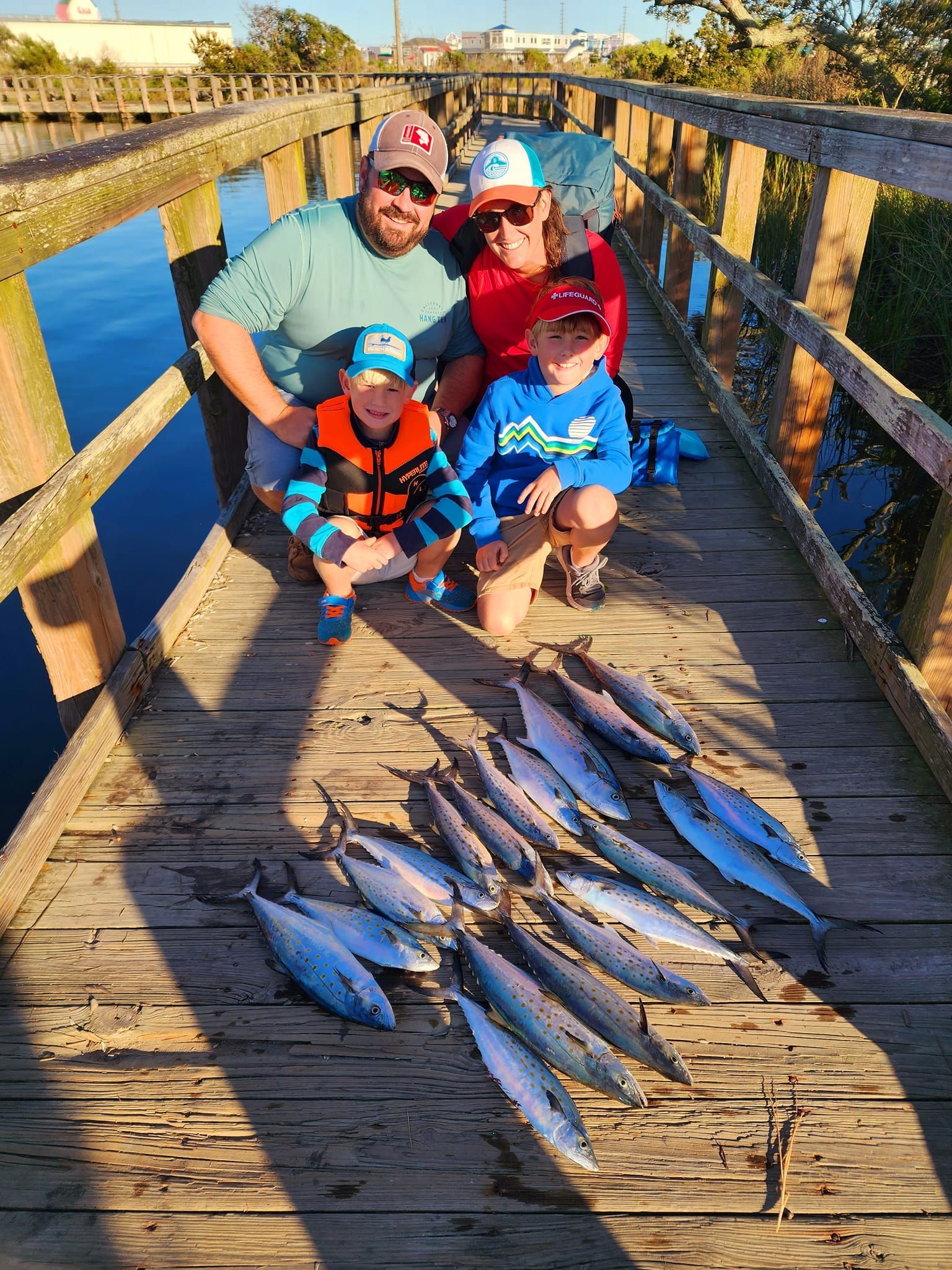 Family on a wooden pier with a pile of caught fish, smiling at the camera. Sunny day, blue water.